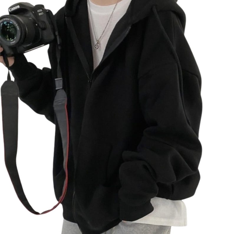 May include: A black zip-up hoodie with a hood and front pockets. The hoodie is worn over a white t-shirt. A black camera with a strap is held in the left hand.