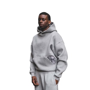 Puede incluir: Una sudadera con capucha gris con un bolsillo de canguro y un pantal&oacute;n de ch&aacute;ndal gris a juego. La sudadera con capucha presenta un parche circular en el bolsillo con un dise&ntilde;o. El conjunto es de un material suave.