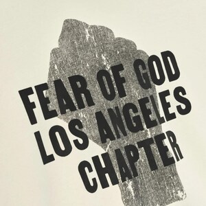 May include: A close-up of a white t-shirt with a black graphic of a clenched fist. The text "FEAR OF GOD LOS ANGELES CHAPTER" is printed in a bold, sans-serif font, angled across the fist.