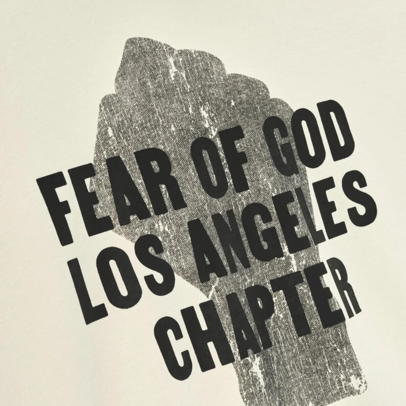 May include: A close-up of a white t-shirt with a black graphic of a clenched fist. The text "FEAR OF GOD LOS ANGELES CHAPTER" is printed in a bold, sans-serif font, angled across the fist.