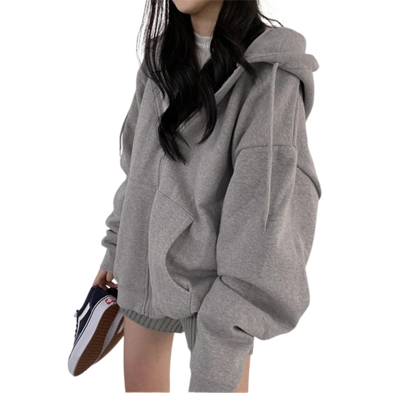 May include: A light gray zip-up hoodie with a hood and large front pockets. The hoodie is made of a soft material and has a relaxed fit. The person is holding a pair of black and white sneakers.
