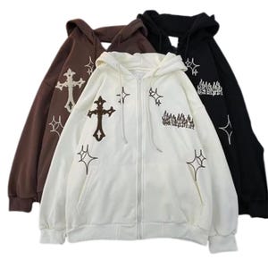 May include: Three hooded zip-up sweatshirts in brown, white, and black. Each sweatshirt features embroidered crosses, starbursts, and text. The white sweatshirt is in the foreground, with the brown and black sweatshirts behind it.