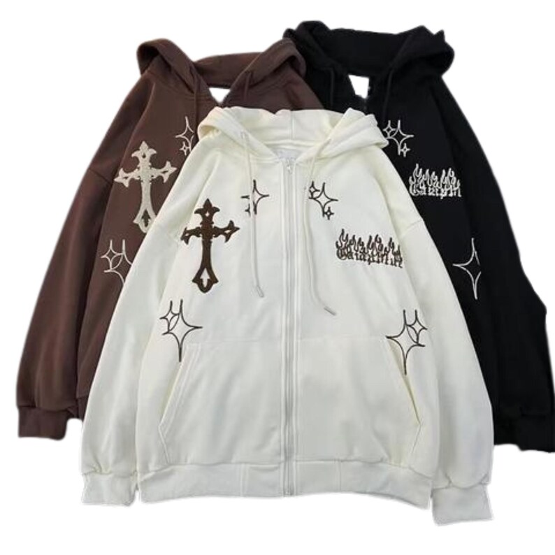 May include: Three hooded zip-up sweatshirts in brown, white, and black. Each sweatshirt features embroidered crosses, starbursts, and text. The white sweatshirt is in the foreground, with the brown and black sweatshirts behind it.