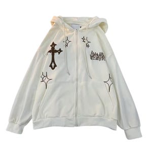 May include: Cream-colored zip-up hoodie with a hood and front pockets. The hoodie features brown embroidered designs, including a cross, stars, and text. The jacket has a full-length zipper and long sleeves.