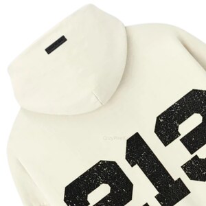 May include: Cream-colored hoodie with a hood and large black numbers on the front. The hoodie has a small black rectangular tag on the hood. The numbers have a distressed, textured appearance.