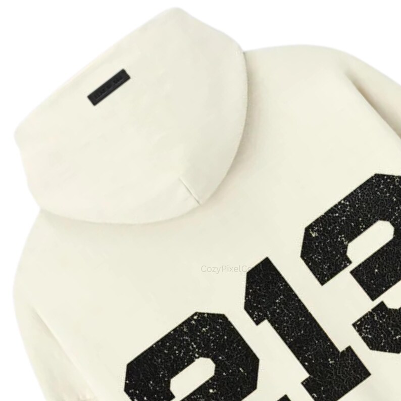 May include: Cream-colored hoodie with a hood and large black numbers on the front. The hoodie has a small black rectangular tag on the hood. The numbers have a distressed, textured appearance.