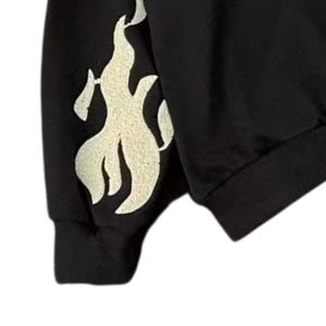 May include: Black sweatshirt with a cream-colored flame design on the sleeve. The flame graphic is stylized and appears to be screen-printed. The sweatshirt has a ribbed cuff and a crew neck.