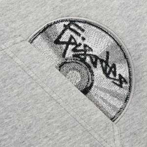Puede incluir: Primer plano de una sudadera gris con un parche en forma de semic&iacute;rculo. El parche presenta un dise&ntilde;o negro y plateado con texto estilizado y un elemento circular, posiblemente un logotipo o emblema. La sudadera tiene un dise&ntilde;o sencillo e informal.
