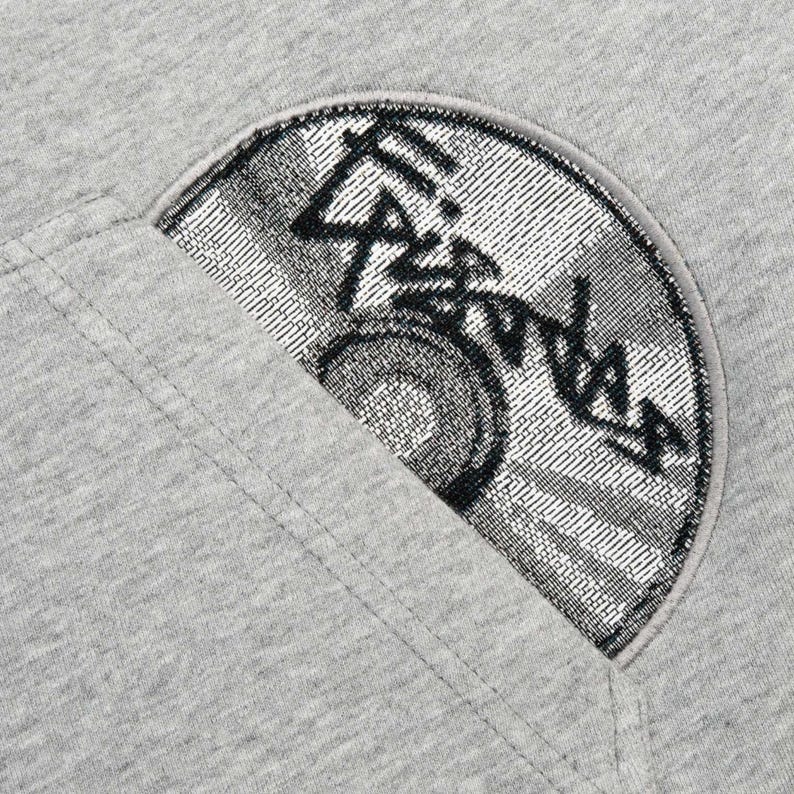 Puede incluir: Primer plano de una sudadera gris con un parche en forma de semic&iacute;rculo. El parche presenta un dise&ntilde;o negro y plateado con texto estilizado y un elemento circular, posiblemente un logotipo o emblema. La sudadera tiene un dise&ntilde;o sencillo e informal.