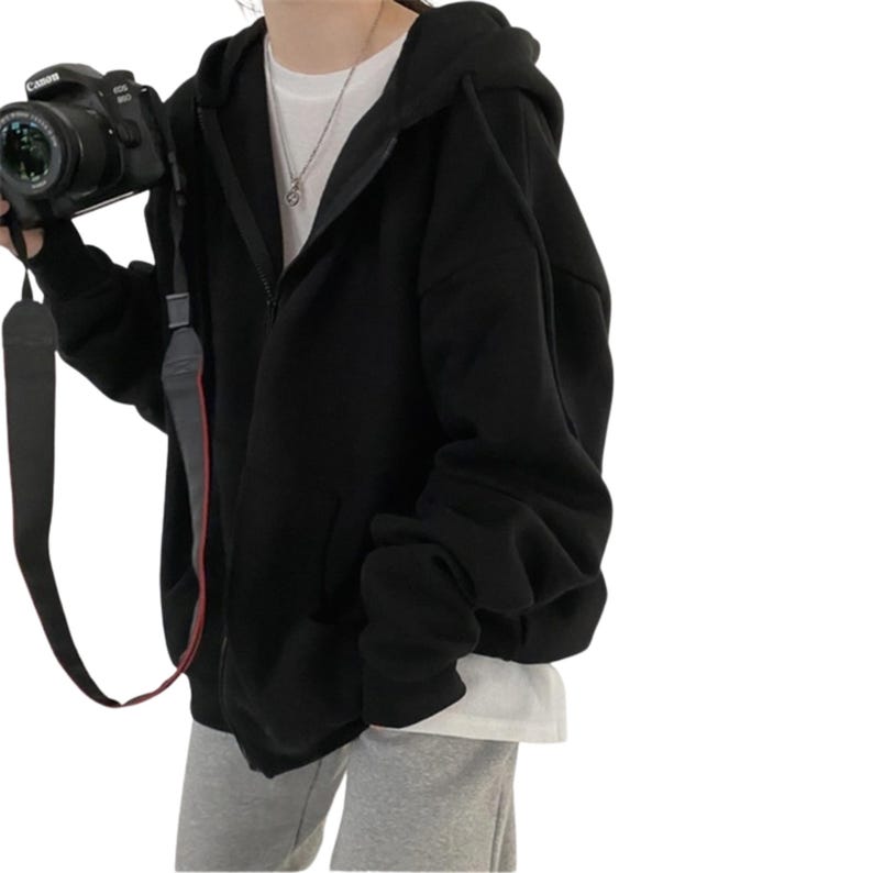 May include: A black zip-up hoodie with a hood and front pockets. The hoodie is worn over a white t-shirt and gray sweatpants. A black Canon camera with a strap is held in the left hand.