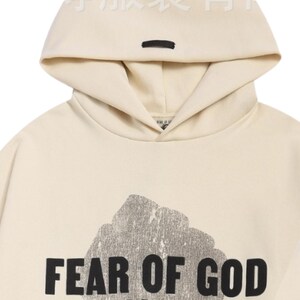 May include: Cream-colored hoodie with a hood and a black rectangular detail. The front of the hoodie features a large, distressed graphic and the words "FEAR OF GOD" in bold, black letters. The hoodie is made of a soft material.