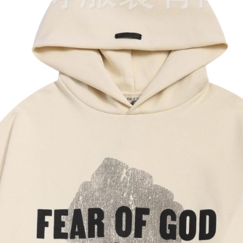 May include: Cream-colored hoodie with a hood and a black rectangular detail. The front of the hoodie features a large, distressed graphic and the words "FEAR OF GOD" in bold, black letters. The hoodie is made of a soft material.