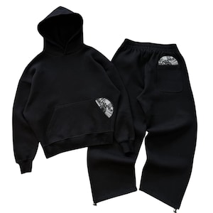 Puede incluir: Conjunto negro de sudadera con capucha y pantal&oacute;n de ch&aacute;ndal. La sudadera tiene un bolsillo delantero y una capucha. Ambas piezas tienen un gr&aacute;fico circular. El pantal&oacute;n tiene cintura con cord&oacute;n y cremalleras en los tobillos.