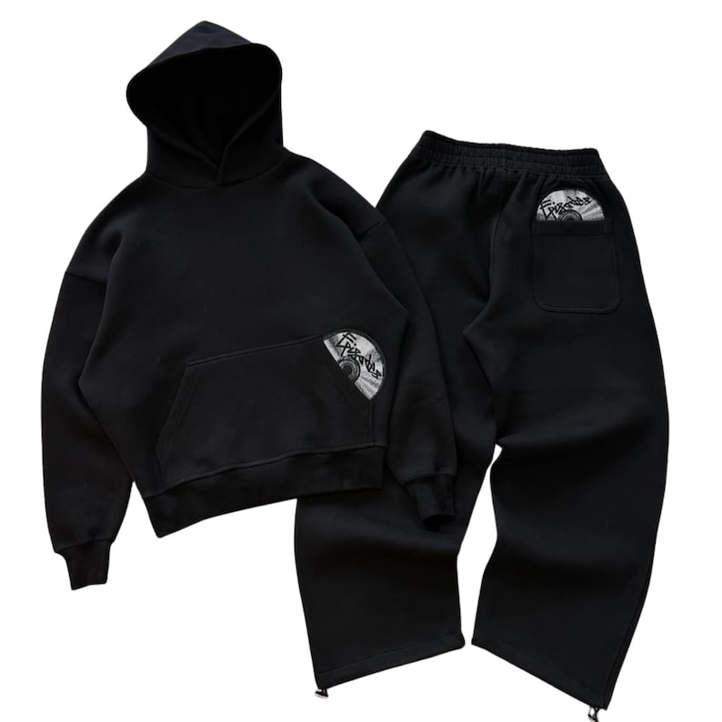 Puede incluir: Conjunto negro de sudadera con capucha y pantal&oacute;n de ch&aacute;ndal. La sudadera tiene un bolsillo delantero y una capucha. Ambas piezas tienen un gr&aacute;fico circular. El pantal&oacute;n tiene cintura con cord&oacute;n y cremalleras en los tobillos.