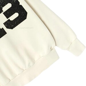 May include: Cream-colored sweatshirt with a large, distressed black number 3 printed on the front. The sleeve and bottom hem have a ribbed texture. The text "CozyPixelCoArt" is printed on the front.