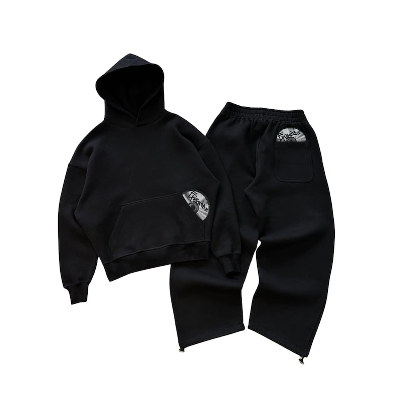 Puede incluir: Conjunto de sudadera con capucha y pantal&oacute;n de ch&aacute;ndal negros. La sudadera tiene un bolsillo delantero y una capucha. Ambas piezas tienen un parche circular con texto y un dise&ntilde;o. El pantal&oacute;n de ch&aacute;ndal tiene un bolsillo trasero.