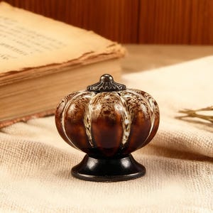May include: A decorative brown and white pumpkin-shaped object with a black base and a decorative top. The object is made of ceramic or glass and is sitting on a beige cloth. The background includes an old book and wooden paneling.