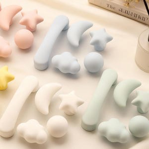 May include: A selection of pastel-coloured cabinet knobs and drawer pulls in various shapes, including moons, stars, clouds, and spheres. The knobs are in shades of pink, yellow, blue, white, and green. The handles are curved and cylindrical.