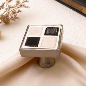 May include: A square cabinet knob with a silver finish and a mosaic tile design. The tile features four squares in black, white, and gray. The knob is mounted on a short, cylindrical base, suitable for furniture hardware.