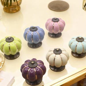 以下が含まれることがあります： 緑、青、ピンク、クリームなど、さまざまなパステルカラーのカラフルなセラミックキャビネットノブのコレクション。 各ノブは、金色のアクセントと濃いブロンズのベースを備えたカボチャのような形をしています。 これらの装飾的なノブは家具用に設計されています。