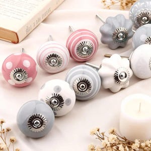 May include: A collection of decorative ceramic cabinet knobs in various colors and patterns. The knobs feature designs such as polka dots and stripes, with silver-toned metal accents. Colors include pink, white, and gray.