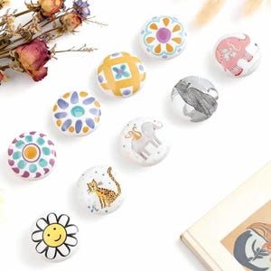 Könnte beinhalten: Eine Sammlung dekorativer Keramikknöpfe mit verschiedenen Designs. Die Knöpfe weisen florale Muster, Tierillustrationen und geometrische Formen in Farben wie Gelb, Rosa, Blau und Weiß auf. Diese Knöpfe eignen sich ideal, um Möbeln eine einzigartige Note zu verleihen.