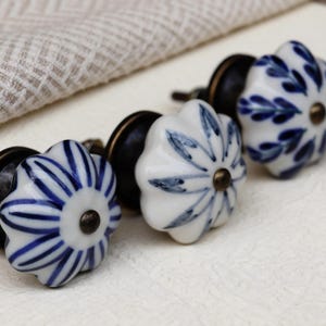 May include: Close-up of three white and blue floral ceramic cabinet knobs with bronze hardware. The knobs have a flower shape with blue painted designs. A neutral-colored cloth is in the background.