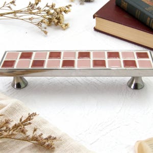 May include: A rectangular, silver-toned serving tray with a mosaic tile design. The tiles alternate between light and dark pink squares. The tray has two decorative, pedestal-style feet. Dried flowers and books are in the background.