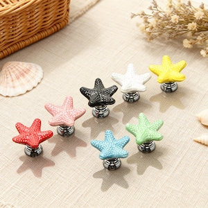 Op de afbeelding: Een verzameling zeestervormige kastknoppen in verschillende kleuren, waaronder rood, roze, zwart, wit, geel, blauw en groen. Elke knop heeft een textuur en een zilverkleurige basis. De knoppen zijn gerangschikt op een neutrale ondergrond.