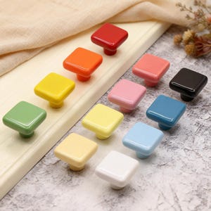 Pode incluir: Uma coleção de puxadores quadrados coloridos para armários em várias tonalidades, incluindo vermelho, laranja, amarelo, verde, rosa, preto, azul e branco. Cada puxador tem uma base arredondada e um acabamento brilhante.