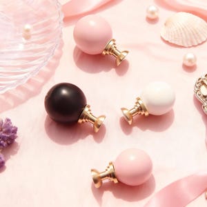 Peut inclure: Quatre bouchons de bouteille décoratifs avec un dessus sphérique rose, noir et blanc, et une base dorée. Les bouchons sont disposés sur une surface rose avec des perles, un coquillage et un ruban.