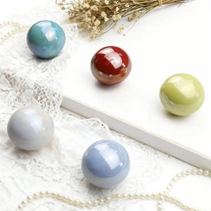 Könnte beinhalten: Fünf schillernde Kugeln in Blau-, Rot-, Grün- und Weißtönen sind auf einer weißen Oberfläche mit Spitze und Perlen angeordnet. Die Kugeln haben eine glänzende Oberfläche und reflektieren das Licht, wodurch ein schimmernder Effekt entsteht.