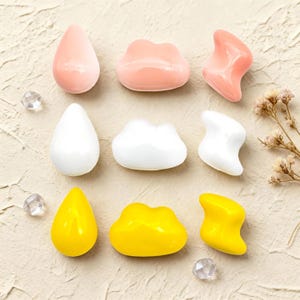 Può includere: Nove oggetti colorati e lisci simili a pietre, di varie forme, tra cui gocce e forme di nuvole. I colori sono rosa, bianco e giallo. Gli oggetti sono disposti su una superficie strutturata di colore chiaro, con piccoli accenti di cristallo.