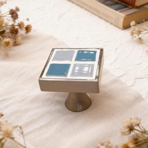 May include: A square cabinet knob with a silver base and a top featuring four blue and gray tiles. The tiles are arranged in a grid pattern, with a silver border. The knob has a pedestal base.