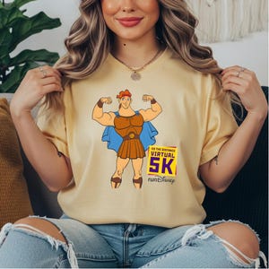 May include: A light yellow t-shirt featuring a cartoon illustration of Hercules flexing his muscles. The design includes a blue cape, brown armor, and the text "GO THE DISTANCE VIRTUAL 5K runDisney" in purple and yellow.