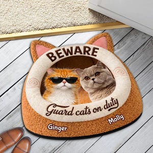 May include: A cat bed shaped like a cat's head, with the text "BEWARE" at the top. Inside are two cats, one wearing sunglasses. The text "Guard cats on duty" is below the cats, with the names "Ginger" and "Molly" below.