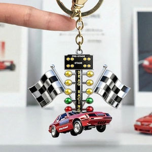 May include: A red drag racing keychain featuring a red car with the name "Michael" on it, a drag racing Christmas tree, and two checkered flags. The keychain is attached to a gold-colored chain.