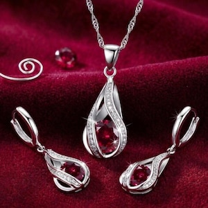 Puede incluir: Un conjunto de joyería compuesto por un collar plateado con un colgante en forma de lágrima y pendientes a juego. Cada pieza presenta una piedra preciosa roja oscura central rodeada por un marco de pequeñas piedras claras y brillantes.