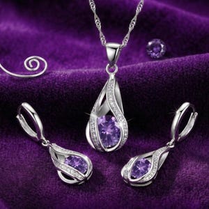 May include: A silver-toned jewelry set consisting of a pendant necklace and two matching drop earrings. Each piece features a central oval-cut purple gemstone encased in a teardrop-shaped frame accented with small clear stones on a silver chain.