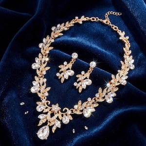 Op de afbeelding: Een goudkleurige ketting en bijpassende oorbellen met een bladvormig ontwerp. De sieraden zijn versierd met heldere, gefacetteerde kristallen in diverse vormen, waaronder marquise- en peervormige slijpvormen, gerangschikt in een rankachtig patroon.