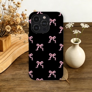 Könnte beinhalten: Schwarze Smartphone-Hülle mit einem Muster aus rosa Schleifen. Die Schleifen sind gleichmäßig verteilt und erzeugen ein zartes Design. Die Hülle hat eine glänzende Oberfläche und ist für ein Smartphone konzipiert.