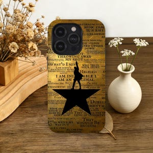 May include: A phone case featuring a design inspired by the musical Hamilton. The case has a gold background with song lyrics and a black silhouette of a character on a star. The case also has the phone's camera and flash.