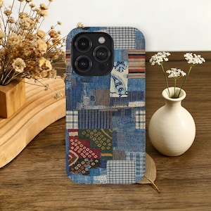 May include: A patchwork-style phone case featuring a mix of blue denim and patterned fabric squares. The case has a camera cutout and is displayed on a wooden surface with a small vase of white flowers and dried plants.