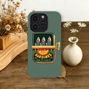 May include: A green phone case featuring a sardine can design. The can is open, displaying three fish with detailed eyes and scales. The label reads "Sardines Pour Les Toasts" in a decorative font, with a red and yellow background.