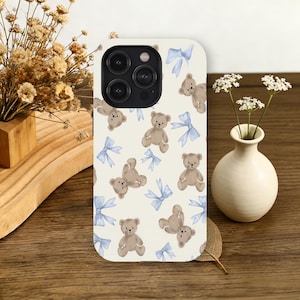 May include: A phone case with a cream background featuring a pattern of brown teddy bears and light blue bows. The case has a rounded rectangle shape with a camera cutout.