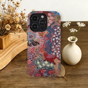 May include: A patterned phone case with a colourful, abstract design. The case features a mix of red, blue, and pink patterns, with floral and geometric elements. The phone case is shown on a wooden surface, with a small vase of white flowers.