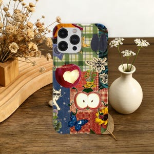 May include: A colorful phone case with a patchwork design featuring an apple with a heart cutout, a cat wearing sunglasses, bees, and a butterfly. The case has a green plaid pattern and a sunflower.
