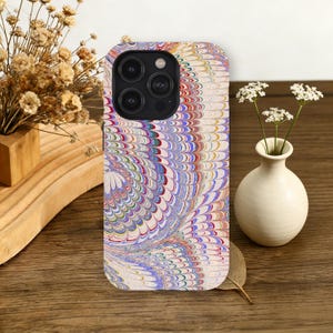 May include: A colorful phone case with a swirling pattern in shades of blue, red, yellow, and white. The case has a camera cutout and is displayed on a wooden surface. A small white vase with flowers is also present.