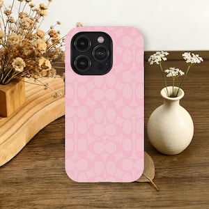 Puede incluir: Una funda de teléfono rosa con un patrón repetitivo de formas ovaladas entrelazadas. La funda tiene un módulo de cámara con tres lentes y un flash. Un pequeño jarrón blanco con flores y una bandeja de madera están en el fondo.