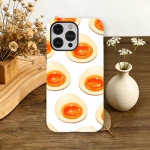 May include: White phone case featuring a repeating pattern of watercolor-style halved soft-boiled eggs. The eggs have a creamy white exterior and a vibrant orange yolk. The phone case has a black camera module.